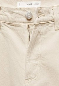 Los pantalones de mezclilla beige presentan un cierre de botón metálico, cremallera y un acabado sutilmente texturizado con costuras en contraste y detalles en los bolsillos.