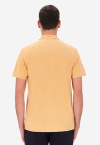 Uomo di spalle che indossa una polo a maniche corte di un leggero colore arancione e pantaloni scuri su uno sfondo bianco semplice.