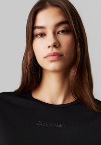 Joven con cabello castaño lacio que lleva una blusa negra de Calvin Klein, expresión neutra, fondo gris liso.