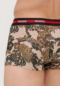 Boxer-briefs floraux avec une base beige ornée de motifs botaniques verts et orange, taille noire avec le logo "HUGO" en rouge.