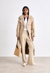 Manteau beige à double boutonnage, avec un large col et un motif texturé ; haut blanc, pantalon beige et sac à main fauve.