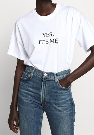 Person iført en hvid t-shirt med teksten „"YES, IT'S ME"“ i sort, kombineret med højtaljede blå jeans, den ene hånd i lommen.