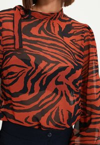 Punainen ja musta läpikuultava pusero, jossa on tiikeriraidallinen kuvio, korkea pääntie ja rusetti kauluksessa. Pitkät, hieman puffatut hihat.