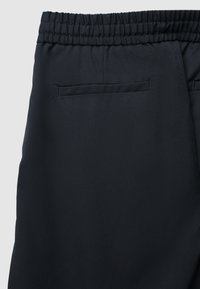 Schwarze, elastische Hose mit elastischem Bund, die eine glatte Textur und eine einzige Gesäßtasche aufweist. Der Stoff hat einen dezenten Glanz und ein strukturiertes Design.