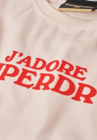 T-shirt di cotone rosa chiaro con testo rosso intenso "J'ADORE SUPERDRY" in un carattere stilizzato e di grande impatto. Tessuto morbido, collo a giroclassico.
