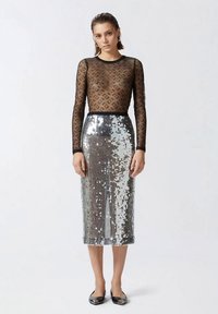 Žena oblečená v čiernom priehľadnom topu s dlhými rukávmi a vzorom, doplnená o striebornú midi sukňu s flitrami a vysokým pásom, stojaca na neutrálnom pozadí.