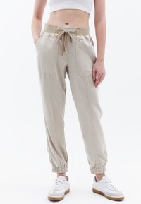 Lichtbeige katoenen joggers met elastische boorden en een trekkoord in de taille, met twee voorzakken en een losse pasvorm. G gedragen met witte sneakers.