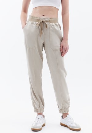 Jogging en coton beige clair avec des poignets élastiques et une taille à cordon, comportant deux poches avant et une coupe décontractée. Porté avec des baskets blanches.