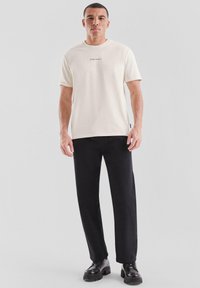 Crème-coloured T-shirt met korte mouwen en een minimalistisch logo op de borst, gecombineerd met zwarte losse broek en zwarte schoenen met een dikke zool.
