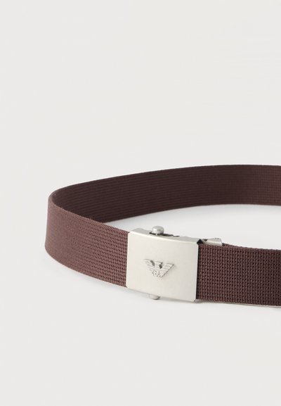 Καφέ υφασμάτινη ζώνη με ασημένια μεταλλική αγκράφα που διαθέτει ανάγλυφο λογότυπο αετού σε απλό λευκό φόντο.