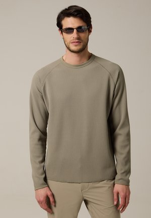 Homme portant des lunettes de soleil foncées, une chemise à manches longues beige texturée et un pantalon beige clair, debout devant un fond neutre uni.