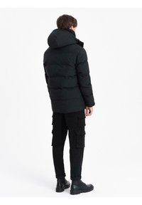 Uomo che indossa un piumino nero con cappuccio, pantaloni cargo neri e stivali neri, in piedi di spalle contro uno sfondo bianco uniforme.