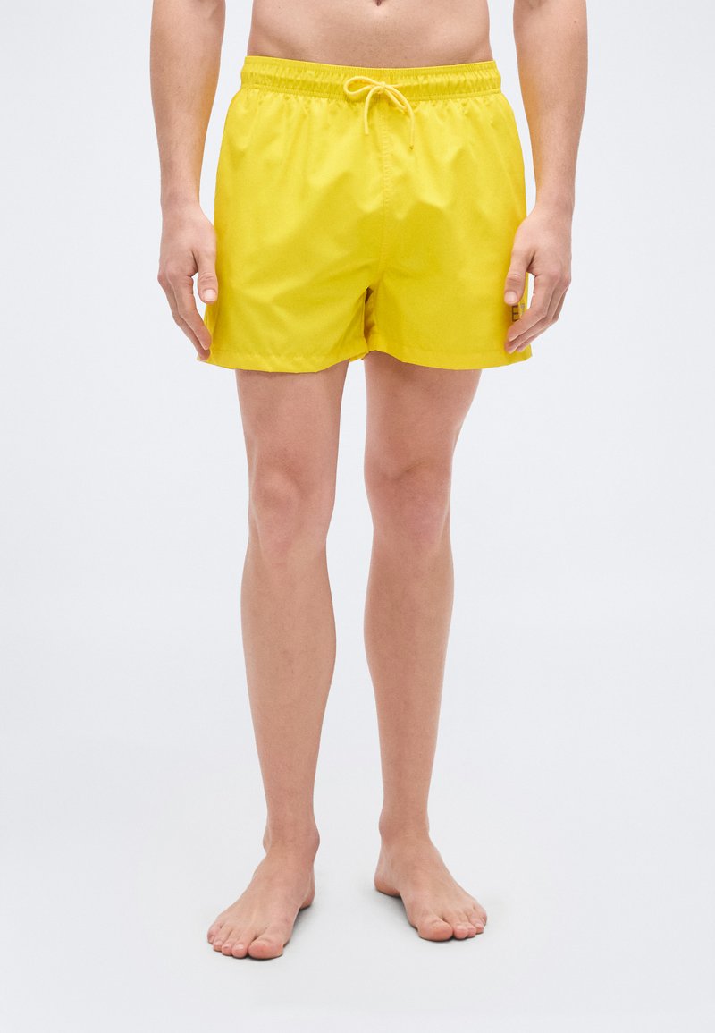 Κίτρινο μαγιό σορτς με ελαστική μέση και ρυθμιζόμενο κορδόνι. Λείο ύφασμα με ελαφριά, γρήγορα στεγνώνουσα υφή. Χωρίς τσέπες.