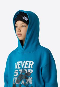 Turkizna jopa z veliko belo in črno grafiko z napisom "NEVER STOP EXPLORING." Ima prostorno kapuco in mehko teksturo. Črna pletena kapa.