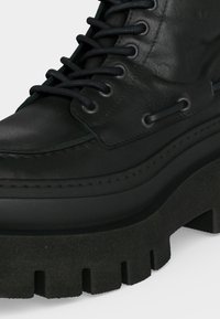 Botte en cuir noire avec une semelle épaisse et texturée, six œillets avec des lacets noirs, et des détails de couture le long de l'avant et des côtés.