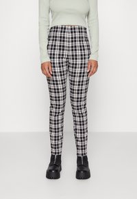 Personne portant un pantalon à carreaux noir et blanc taille haute, un haut à manches longues vert clair, et des bottes noires à semelle épaisse, debout sur un sol blanc.