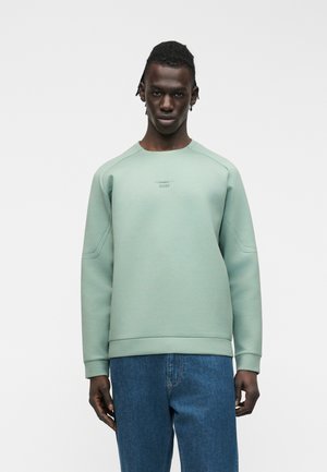 Muž v svetlozelenom pulóvri a modrých džínsach stojaci pred jednofarebným bielym pozadím, tvárou smerujúci dopredu s neutrálnym výrazom.