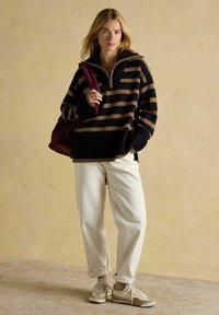 Gestreifter Pullover in Schwarz und Braun mit einem halben Reißverschluss-Kragen, kombiniert mit weißen Hosen und beigen Sneakers. Der Hintergrund hat einen sanften, neutralen Farbton.