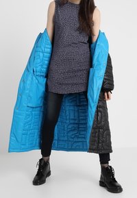 Long black and blue quilted coat with a shiny finish, featuring large pockets and button closures, worn over a dark patterned sleeveless dress.