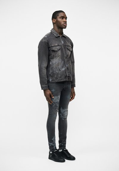 Άνδρας όρθιος, φοράει σκούρο τζιν μπουφάν πλυμένο και τυπωμένο skinny τζιν με μαύρα αθλητικά παπούτσια μπροστά σε λευκό φόντο.