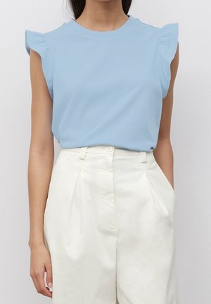 Vrouw die een lichtblauw mouwloos topje met geschulpte schouders en een witte plissébroek met hoge taille draagt, staand tegen een effen achtergrond.