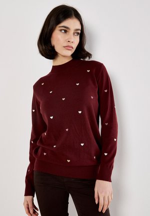 Pull en maille bordeaux avec un col rond, présentant des motifs de cœurs métalliques dispersés, des poignets et un ourlet en côtes. Texture douce, coupe décontractée.