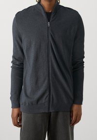 Mörkgrå zip-up tröja med hög krage, tillverkad av stickat material; har långa ärmar och ribbade muddar och fåll. Enkel design utan mönster.