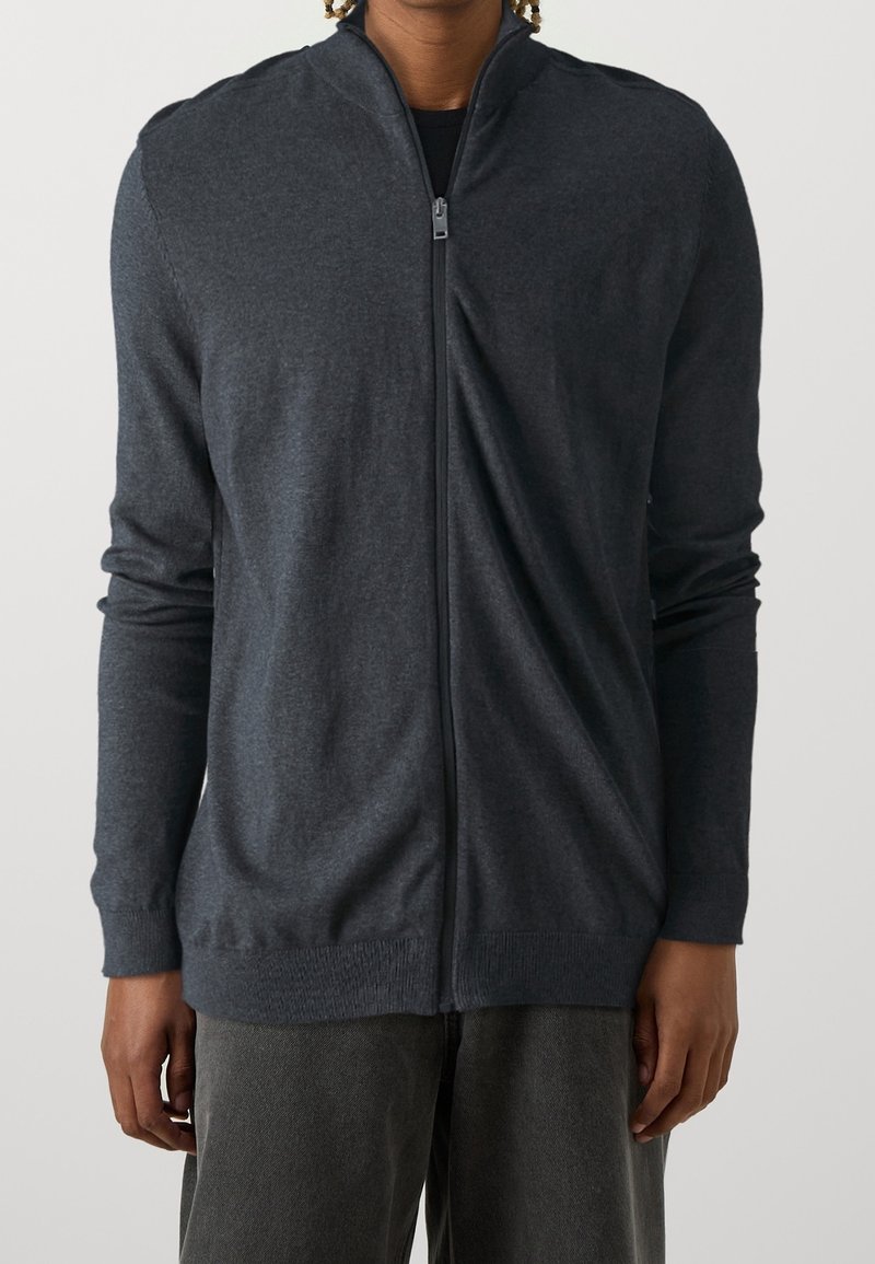 Mörkgrå zip-up tröja med hög krage, tillverkad av stickat material; har långa ärmar och ribbade muddar och fåll. Enkel design utan mönster.