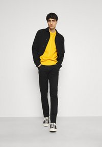 Jachetă neagră, pulover cu tricotaj galben, pantaloni negri și adidași alb-negru. Modelul stă cu mâinile în buzunare pe un fundal neutru.