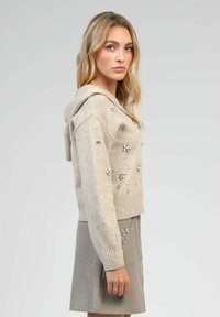 Pull à capuche beige avec un ourlet côtelé, orné de décorations florales en argent. Port�é avec une jupe grise, mettant en valeur un tissu texturé.