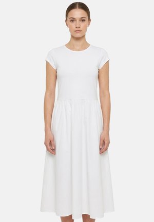 Giovane donna in piedi, di fronte, che indossa un vestito midi bianco a maniche corte con corpino aderente e gonna arricciata, su uno sfondo semplice.