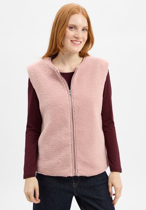 Jonge vrouw met rood haar die een lichtroze fleece vest met rits over een langemouwen donkerburgundy top en donkere jeans draagt, terwijl ze lacht.