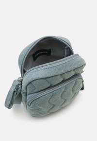 Prešívaná modrá denim taška s dvoma zipsami, ktorá má priestranný interiér a štruktúrovaný povrch. Kompaktný tvar so bočnou rukoväťou.