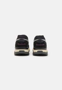 Čierne tenisky s textúrovaným čiernym materiálom na päte, sivými akcentmi a béžovou podrážkou. Na zadnej strane je vyšívané "Reebok" v purpurovej farbe.