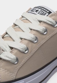 Κοντινό πλάνο μπεζ αθλητικού παπουτσιού Converse All Star με λευκά κορδόνια περασμένα από μεταλλικά μεταλλικά σημεία και ορατό μαύρο και λευκό περίγραμμα στη σόλα.