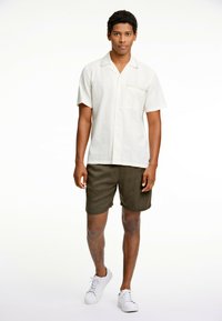 Korte mouwen, crèmekleurig overhemd met een gestructureerd patroon; olijfgroene shorts en witte sneakers maken de outfit compleet. Staande model poseert voor een effen achtergrond.