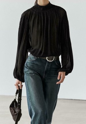 Personne portant un chemisier noir à manches longues et un jean bleu, tenant un petit sac à main noir à ses côtés.