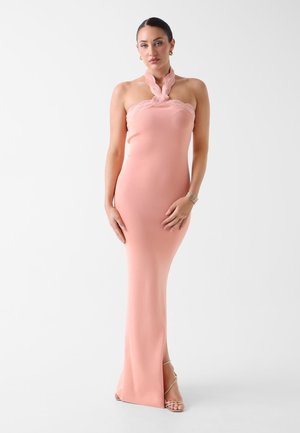 Kobieta w bezramiennych, sięgających podłogi, brzoskwiniowej sukni z zakręconym dekoltem halter i koronkowym obszyciem, stojąca na tle jednolitego jasnego tła.