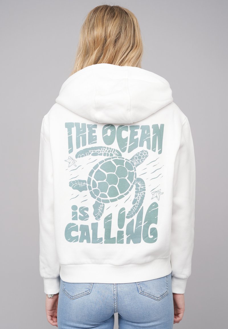 Felpa con cappuccio bianca caratterizzata da una grafica di una tartaruga marina e dal testo "L'OCEANO STA CHIAMANDO" in turchese. Tessuto morbido, vestibilità comoda.