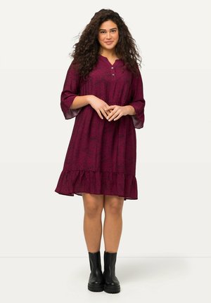 Robe bordeaux avec une jupe évasée, des manches trois-quarts et un décolleté en V. Présente un motif subtil et un ourlet à volants. Portée avec des bottes noires.