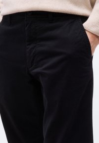 Άτομο που φοράει μαύρο παντελόνι με το δεξί χέρι στην τσέπη, συνδυασμένο με μπεζ πουλόβερ, εμφανιζόμενο από τη μέση μέχρι το μέσο των μηρών.