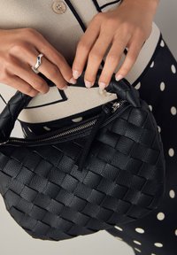 Sac à main en cuir tressé noir avec une forme incurvée, fermeture éclair et anse torsadée. Détail notable de texture et de motif sur la surface.