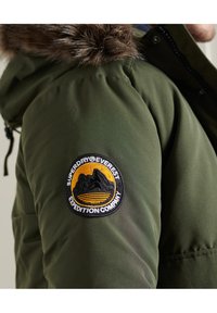 Grøn polstret jakke med en aftagelig faux fur hætte, med et cirkulært sort og gult mærke, der siger "Superdry Everest Expedition Company."