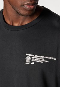 Musta puuvillat-paitaa, jossa on valkoista brodeerausta "Worldwide Hoops", koripallokuvasto ja Adidas-logo rinnassa.
