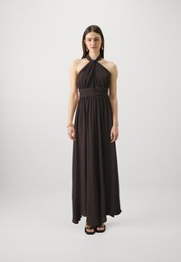 Robe maxi halter marron foncé avec un corsage plissé et des détails à la taille. Tissu léger, longueur au sol, avec une jupe fluide. Portée avec des sandales noires.