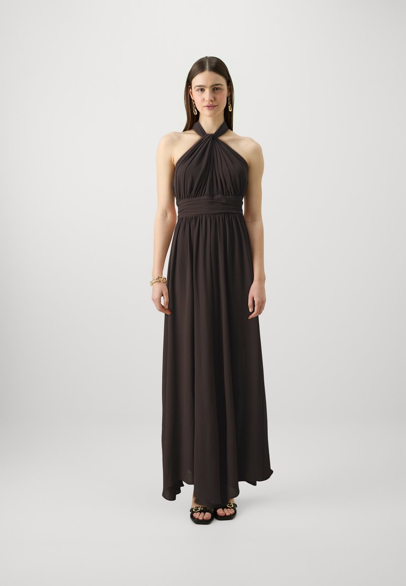 Robe maxi halter marron foncé avec un corsage plissé et des détails à la taille. Tissu léger, longueur au sol, avec une jupe fluide. Portée avec des sandales noires.