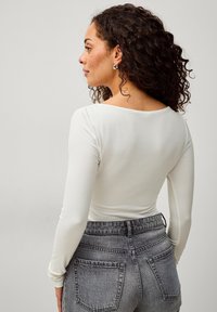 Weißes Langarmoberteil mit einem tiefen Rückendesign, kombiniert mit hoch taillierten grauen Jeans aus Denim mit verwaschener Textur und hinteren Taschen.