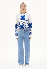 Weißer Strickpullover mit blauem Blumenmuster; kombiniert mit hellblauen, weiten Jeans. Weiße Schuhe runden das Outfit ab.