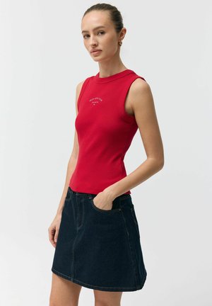 Kobieta ubrana w czerwoną bluzkę bez rękawów z napisem "True Love Club" i ciemną spódnicę dżinsową, pozująca z jedną ręką w kieszeni spódnicy.