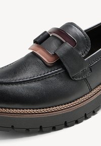 Mocassin en cuir noir avec semelle épaisse et double boucle en métal sur le dessus, avec une couture contrastante marron le long de la semelle.